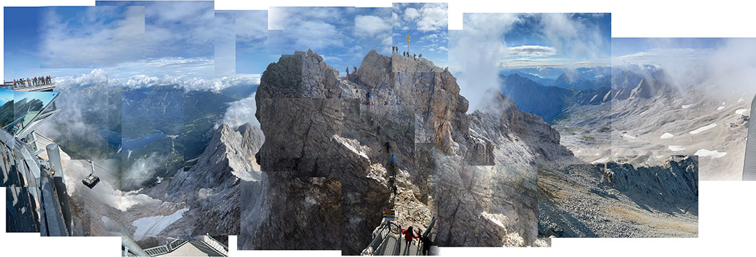 Ein große querformatige Panorama-Abbildung, die sich wie ein Puzzel aus vielen verschiedenen Fotos zusammensetzt mit der Zugspitze in der Mitte. Oben auf dem bergrücken stehen neben dem Gipfelkreuz einige Bergsteigerinnen und Bergsteiger.